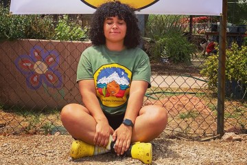 Raquel Gomez sits crossed legged on the ground with her eyes closed smiling at the camera. The sign behind her says Atabey Outdoors.