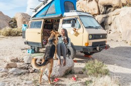 Noami is smiling while her dog jumps up on her lap. She's sitting on a big rock and her yellow Volkswagen van is behind her.