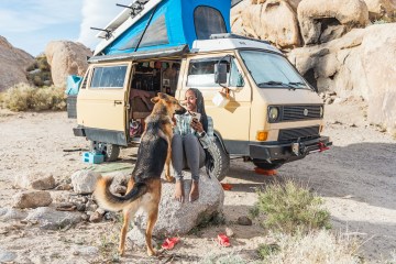 Noami is smiling while her dog jumps up on her lap. She's sitting on a big rock and her yellow Volkswagen van is behind her.