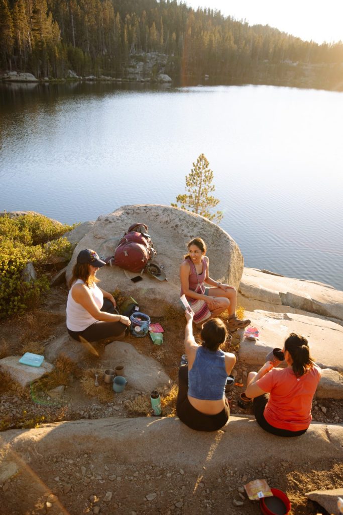 A group of friends isit on a rocky outcropping over a body of water, sharing a meal.