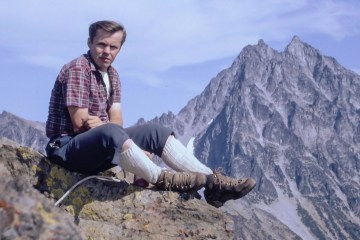 Dave Chantler on Ingalls Peak with blue skies and mountain peaks in the background. He is wearing a short sleave red plaid shirt, blue grey knickers, tall white socks and brown leather mountaineering boots.