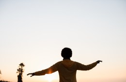 A woman wearing an orange REI Co-op Flash Jacket has her arms out and is silhouetted by the setting sun.