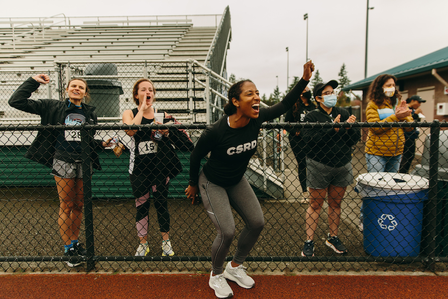 Ashley Davies, co-founder of Club Seattle Runners' Division, stands in front of fence, on a track, cheering on runners. Other runners stand behind her and behind the fence, also cheering.