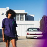 A woman wearing sunglasses, a purple shirt and black compression running shorts stands in front of a white, modern house with a classic white car in the driveway.