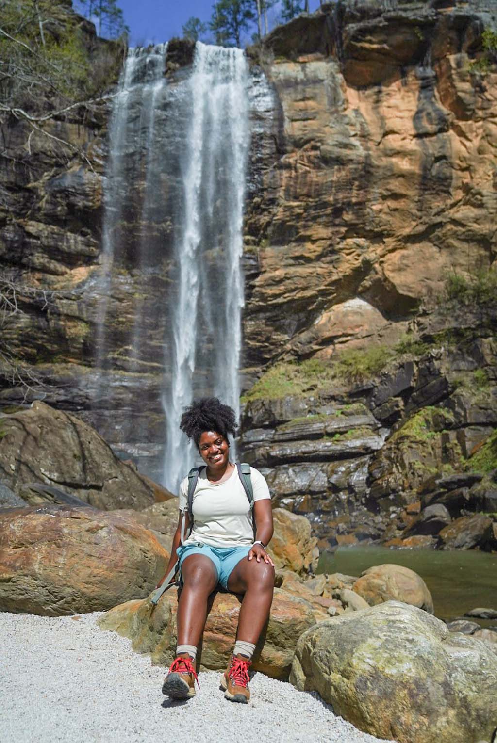 Tee George is smiling and seated in front of a large waterfall. 