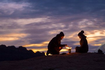 Two campers cooking food over a portable wood-burning camp stove