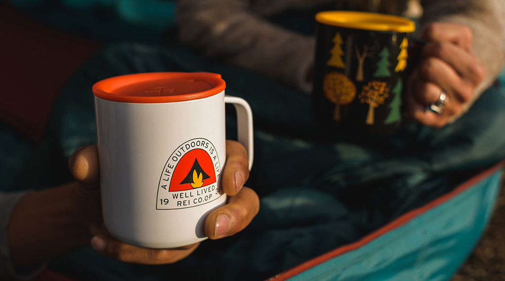 Two people drink from travel mugs while sitting on a sleeping bag