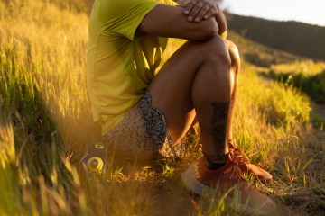 A person sitting on a grassy hill in a pair of red REI Co-op Swiftland MT Trail-Running Shoes.