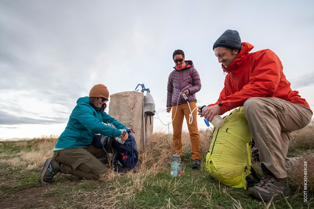 Three people in hiking gear filling up a water bottle from a pump, using their MSR Guardian Gravity Purifier.
