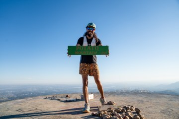 Alan Ortega in running gear on the top of a mountain holding a sign that says "Potato Mountain." There are also several potatoes at his feet.