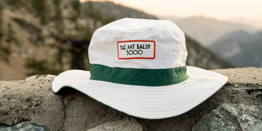 Perched on a mountain ridge, a white bucket hat with a green band around around the base of the cap and the words "THE MT BALDY 5000" written in green with a read square border around the words. On the side of the hat is an illustrated map of Mt. Baldy.