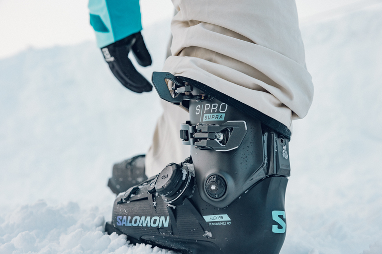 a person's hand reaches down to adjust ski boot that has a BOA dial.