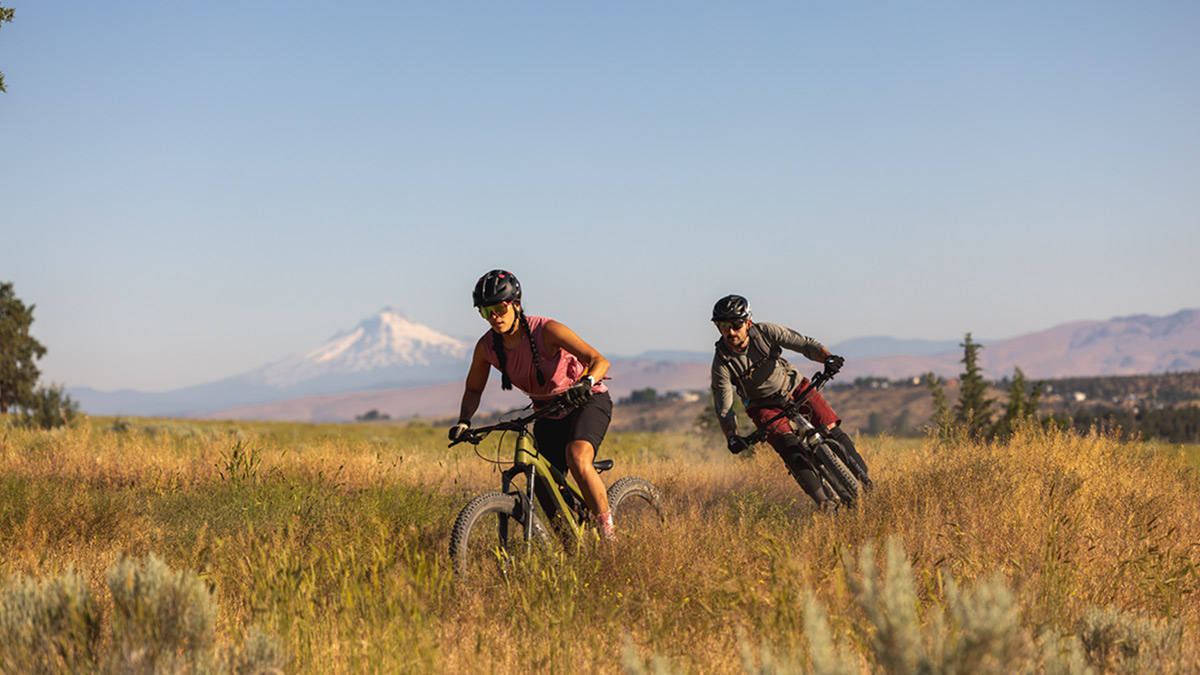 Two cyclists riding in a field against the backdrop of a mountain.
