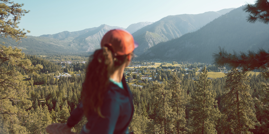 The back of a runner in a red hat and blue jacket. The runner is stretching while standing up and facing a beautiful mountain view. 