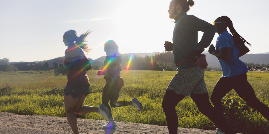 Four people run on a flat trail, with a grassy field alongside the trail. The afternoon sun beams on them.