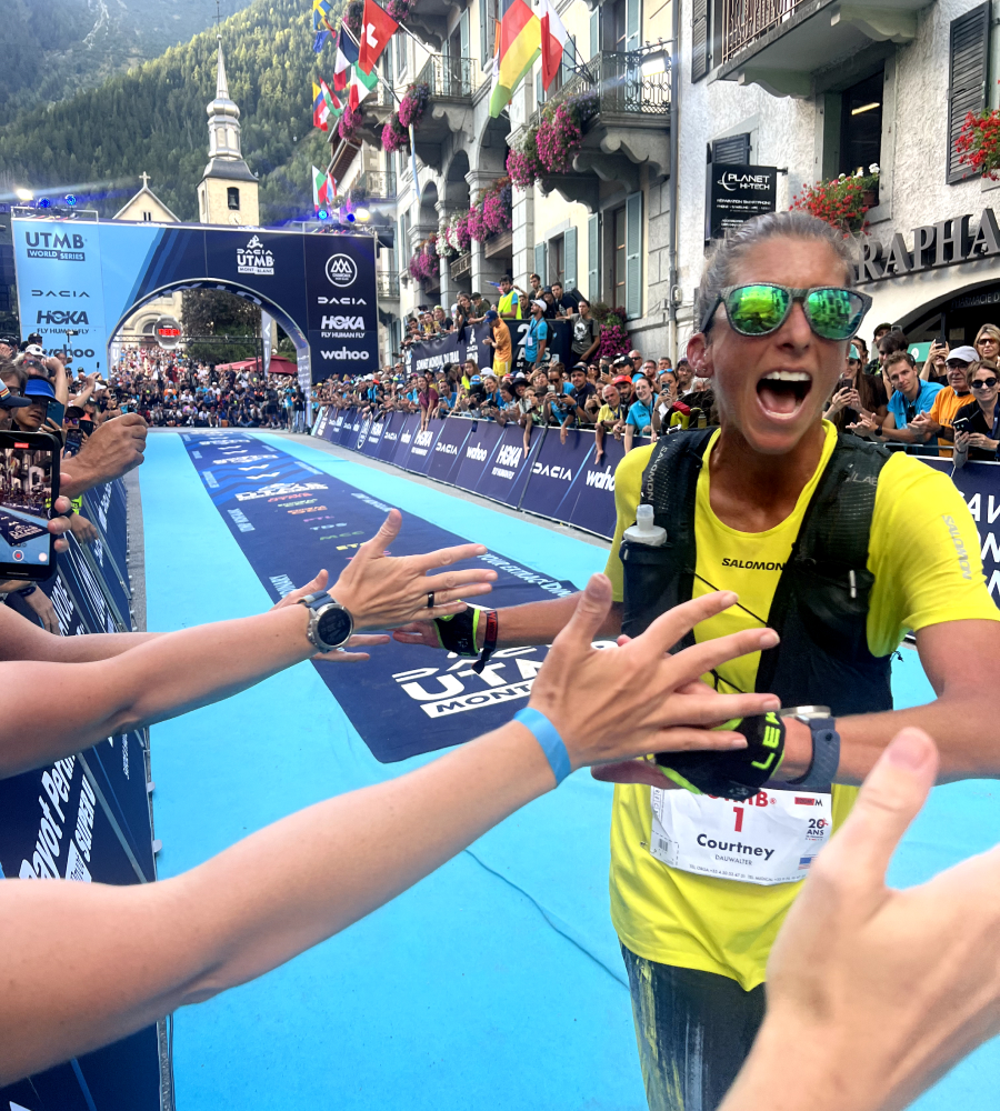 A professional athlete in a yellow technical shirt and black hydration vest high-fives spectators after crossing the finish line.