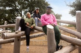 Three people dressed in the REI Co-op Active Pursuits collection, sitting together on a wooden structure in relaxed poses and looking at the camera.