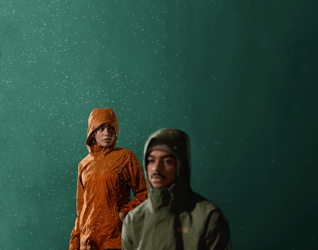 Two people wearing REI Co-op Rainier Rain Jackets stand in wet conditions in front of a dark green backdrop. 