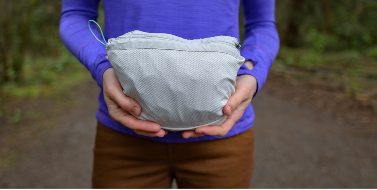 A model, who is wearing a purple long-sleeved shirt and brown pants, holds the Rainier rain jacket, which is folded up into its "packable" pocket.