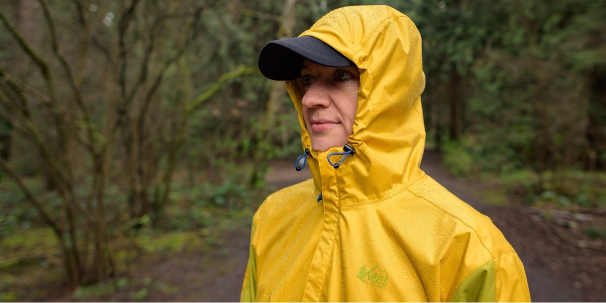 The authors wears the Trailmade rain jacket in Moss Citron/Goldenrod, which is a bright yellow color way, with the hood cinched and a black baseball cap underneath the hood.