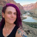 A person with shoulder-length dyed-purple hair looks into the camera for a selfie in front of a river that runs through the base of a canyon.