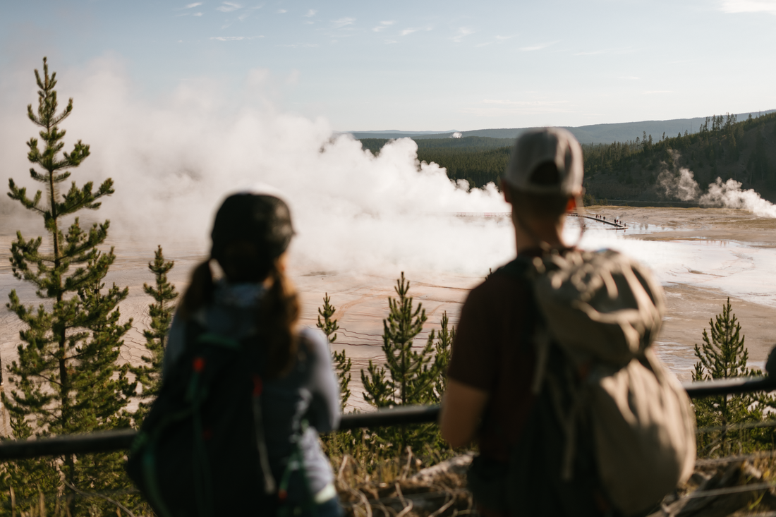 Two hikers wearing packs look at a geyser