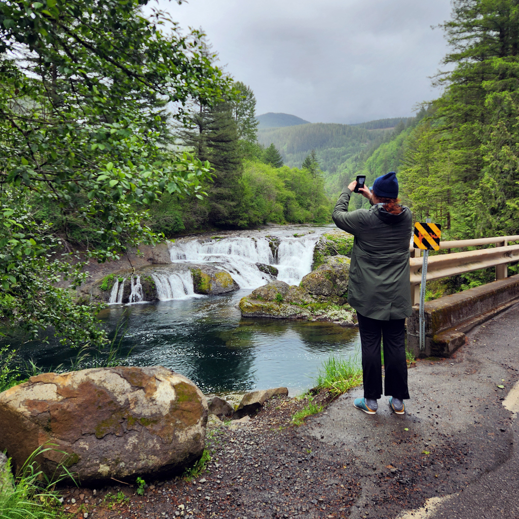 Image of a woman snapping a photo of a small waterfall near the side of a road.