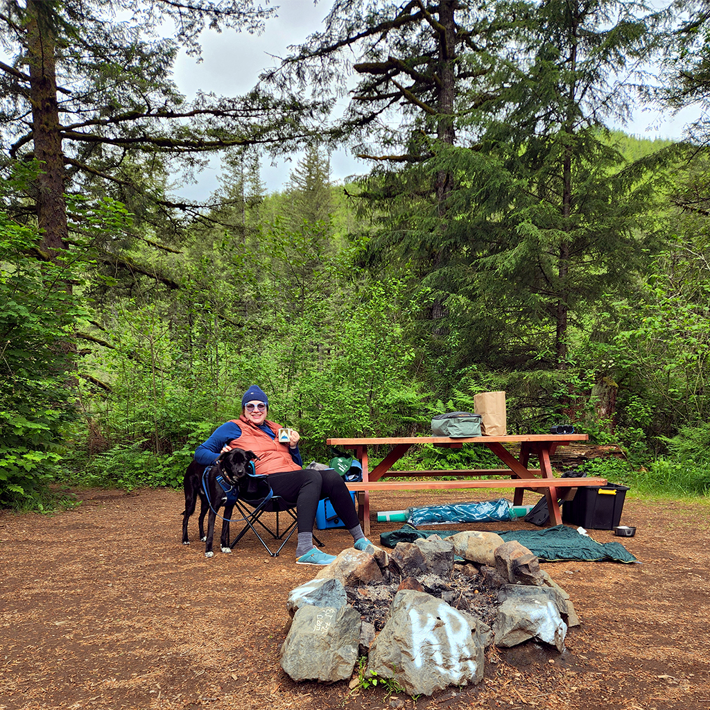 Image of a woman sitting at a picnic table at a campsite. A black dog stands near her.