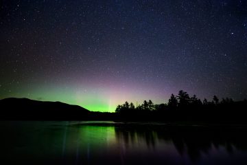 A view of the night sky from across a body of water. The northern lights are glowing green and pink and stars are visible in the sky.