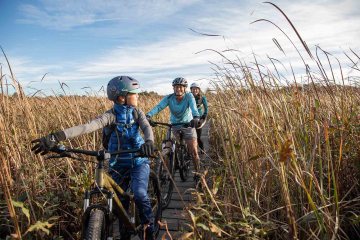 A family is biking along a plank bike trail in tall grass