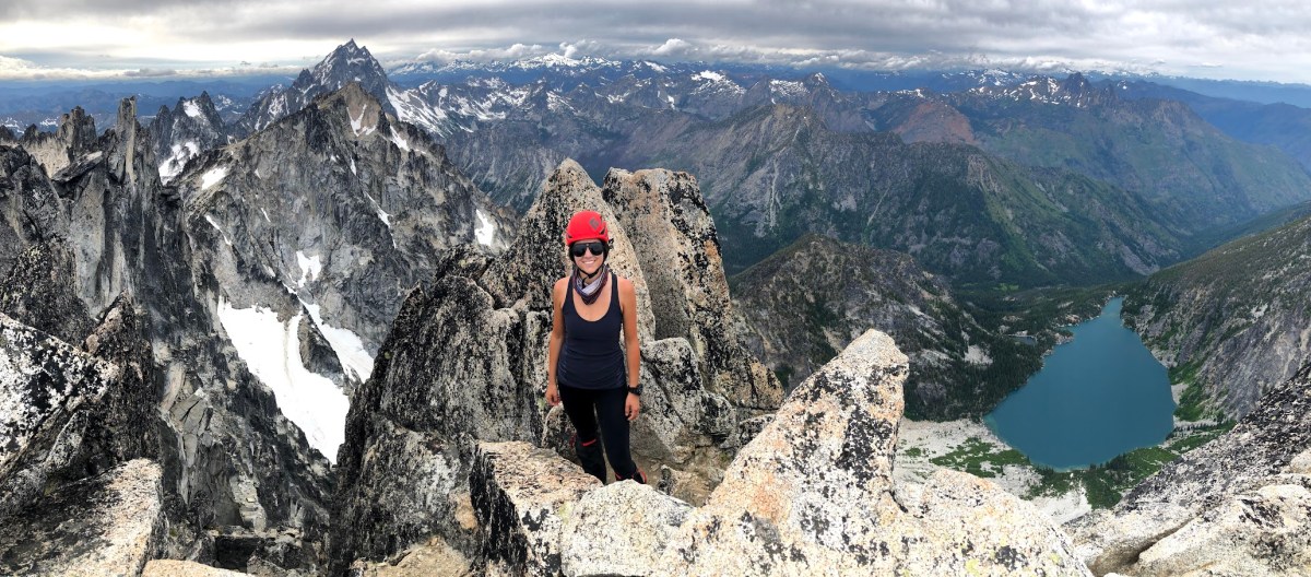 A person stands on the top of a rocky summit with views of snow and an alpine lake below.