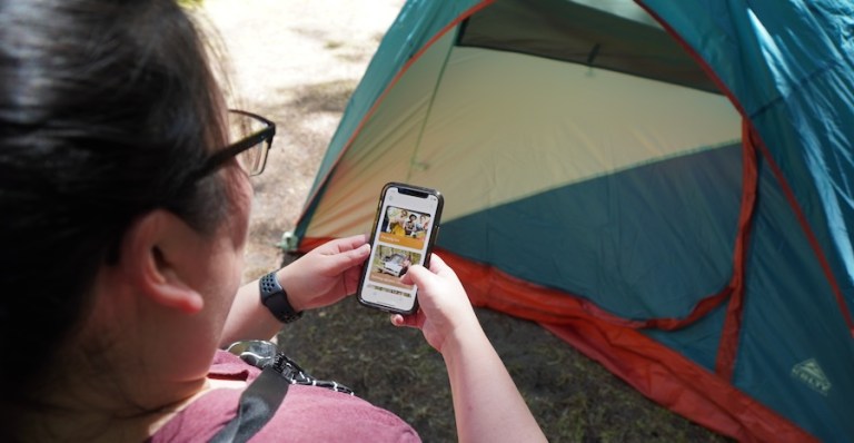 A camper standing in front of a tent uses the Fridie Outdoors app.