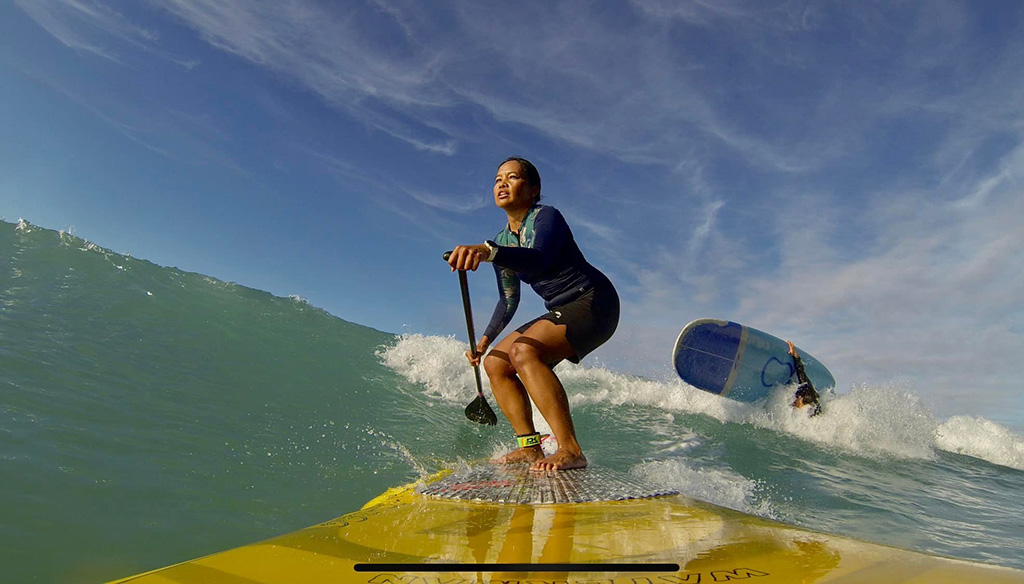 Looking down the end of a surfboard, we see a woman standing up, holding a paddle and riding a wave.