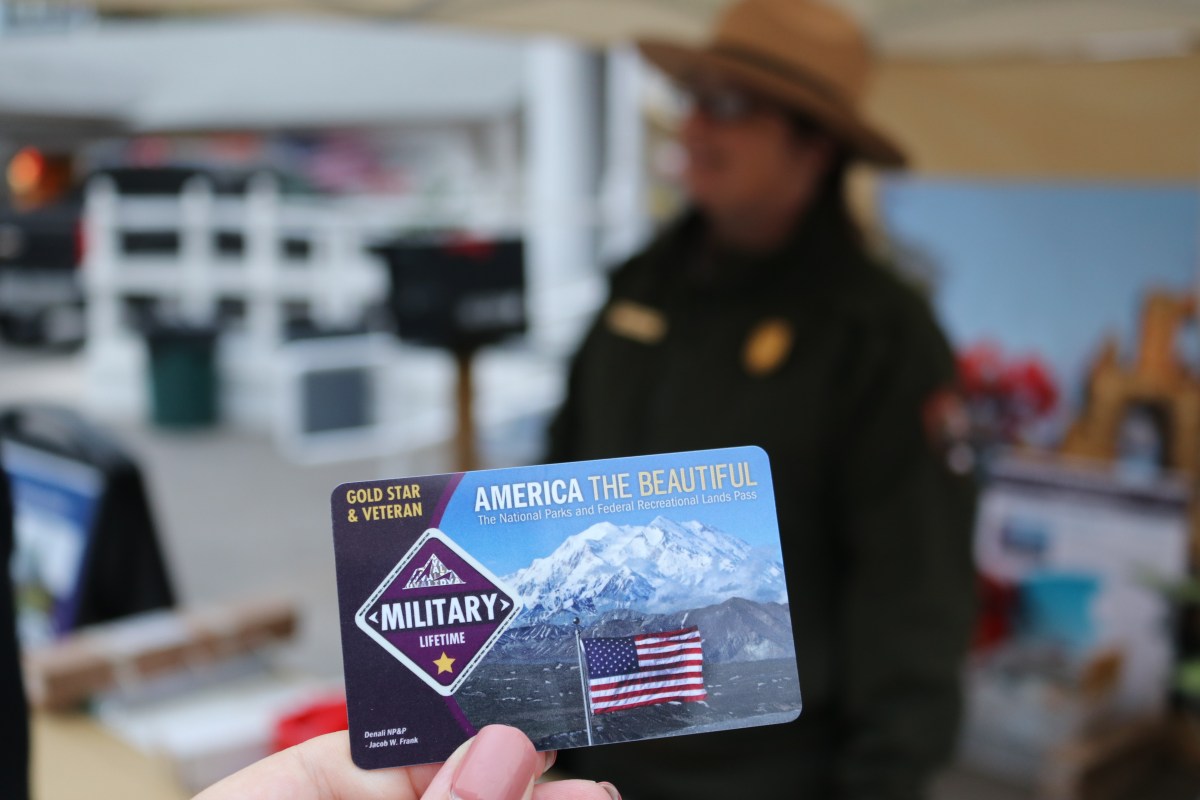 A hand in the foreground holds up an America the Beautiful military lifetime parks pass. 