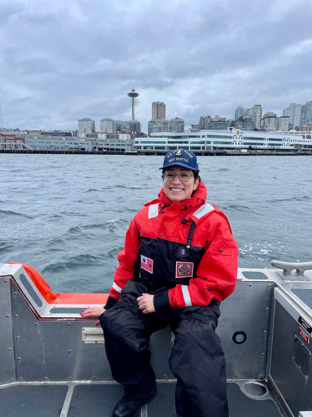 A smiling person wearing a US Coast Guard crew uniform, sitting on a Coast Guard ship in the water