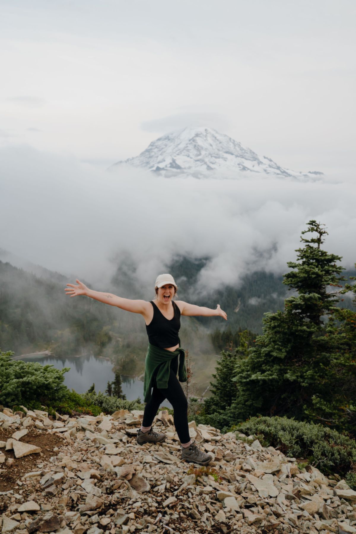 A hiker with a wide smile stands with arms outstretched at the top of a mountain.