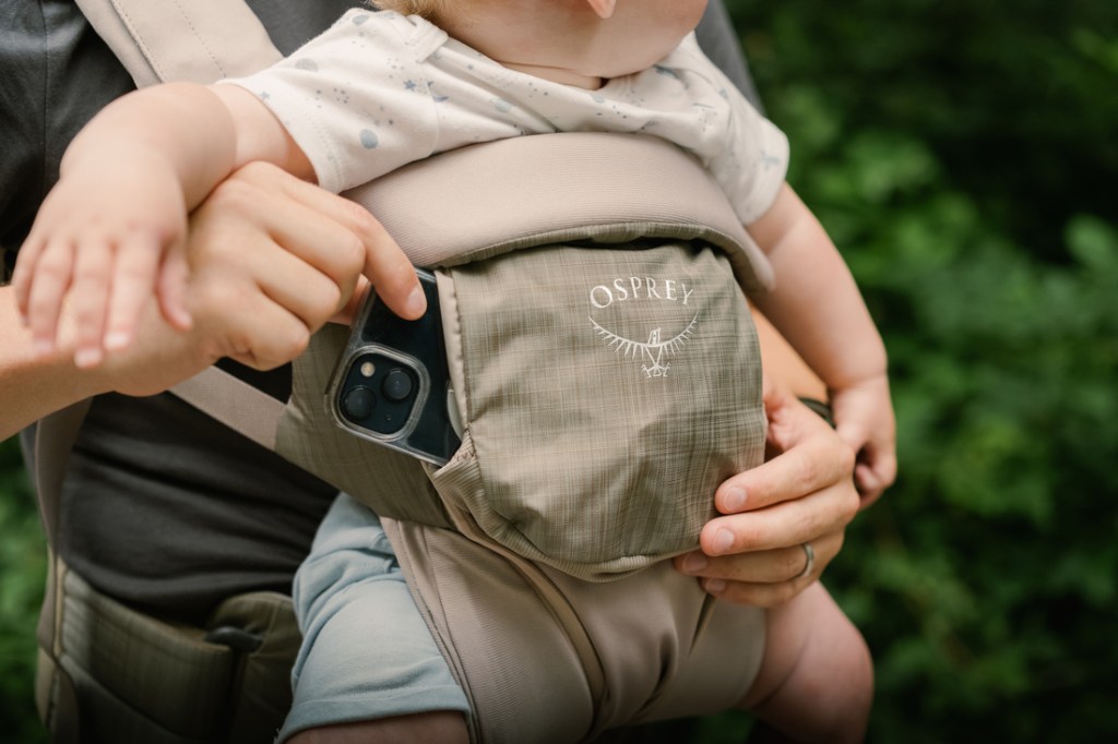 A person wearing a soft baby carrier is shown slipping a smartphone into its pocket.