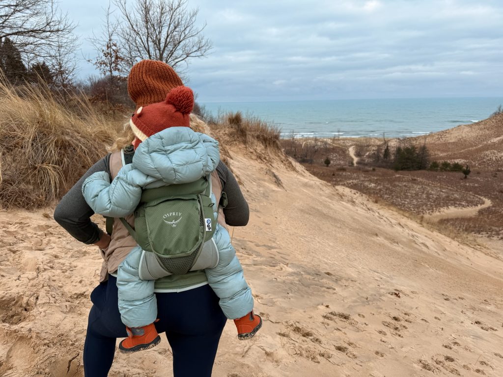 A person wearing a soft child carrier with a toddler in it stands against a dune formation
