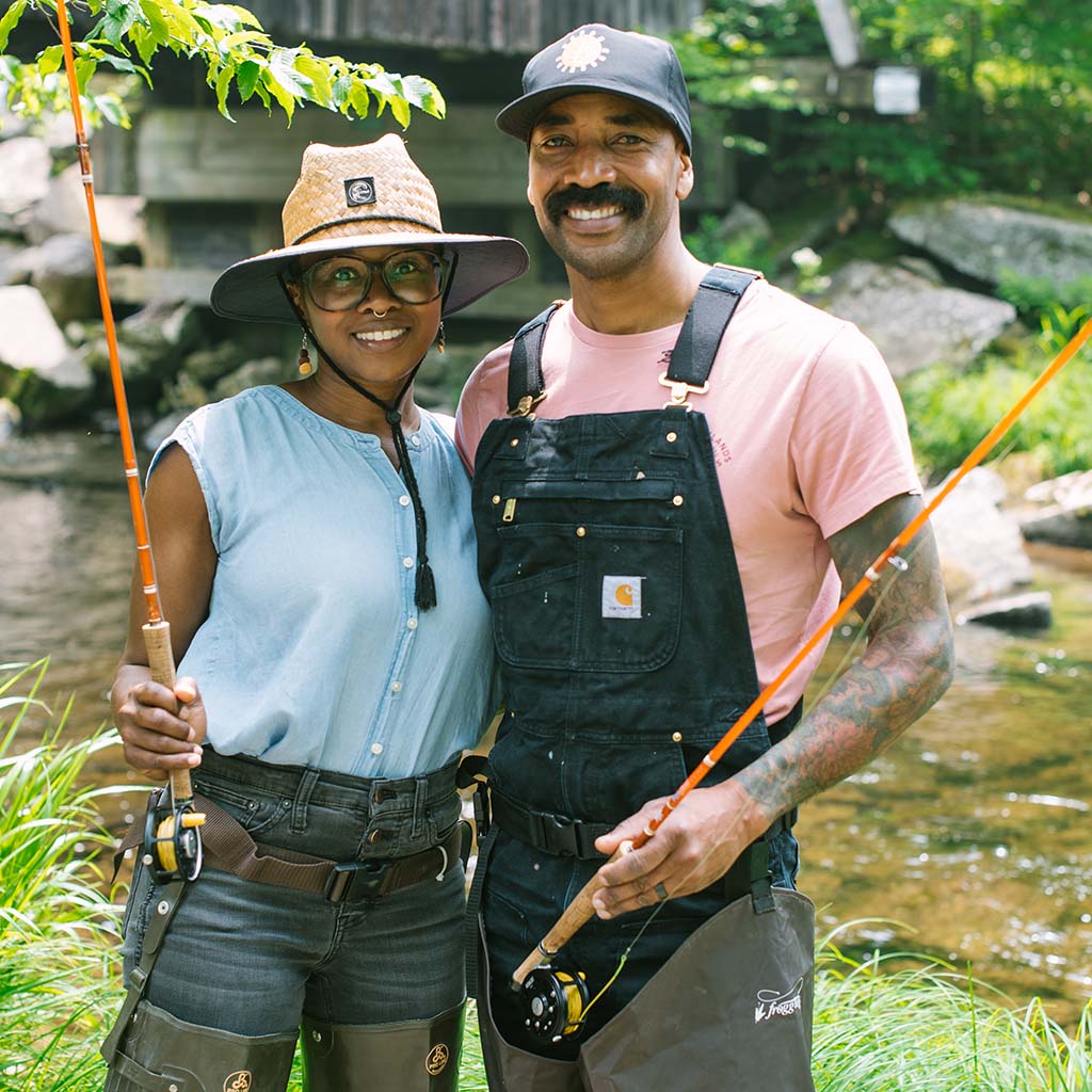 A Black couple stands in front of a water source holding fishing rods