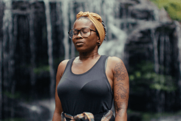 A Black woman wearing glasses, a headwrap and a tank top, standing in front of a waterfall.