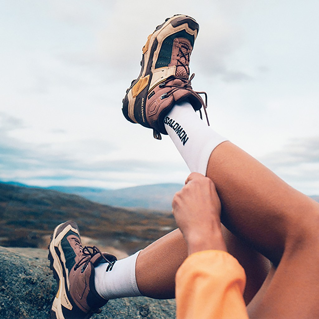A person leaning back raises their left leg in the air to pull up a pair of socks that say Salomon; they are wearing mid-height hiking boots.