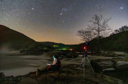 A person sits by a body of water, illuminated by his cell phone light. Next to him is a camera, and above him the sky is full of stars