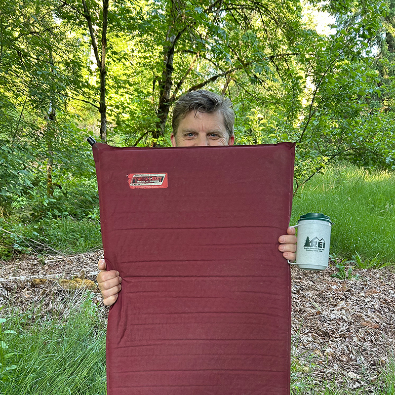 Greg Wayt holds a maroon sleeping pad and classic REI insulated mug