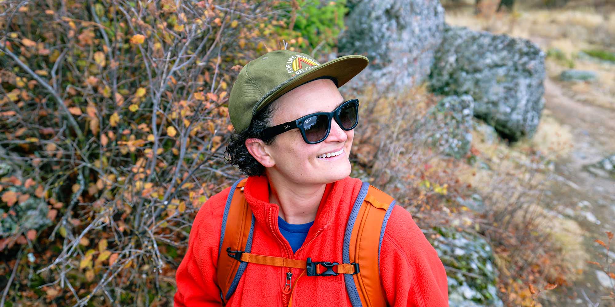 A person wearing an REI Co-op hat, orange fleece jacket and Ray-Ban sunglasses smiles off into the distance.