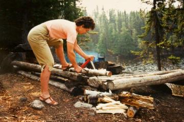 A person in the woods is bent over a downed tree and uses a portable, folding saw.