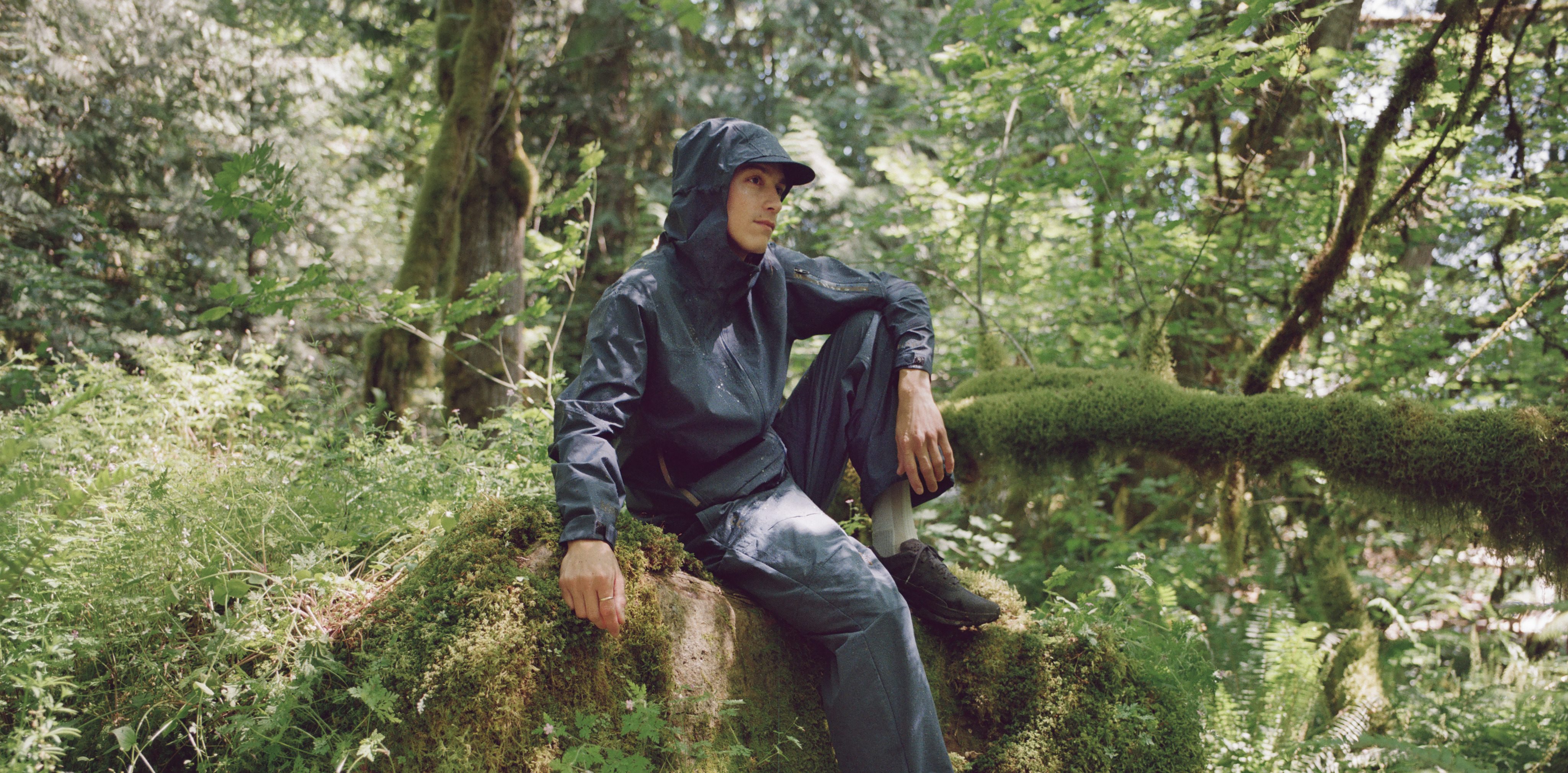 A man in navy-colored rainwear and trail-running shoes sits on a mossy rock in a lush forest.