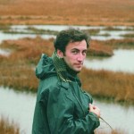 A young man with brown hair standing in front of a marsh, wearing an olive-colored rain jacket. His head is turned towards the camera.