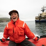 A person wearing a bright orange jacket on a raft in the water laughs with an open mouth.